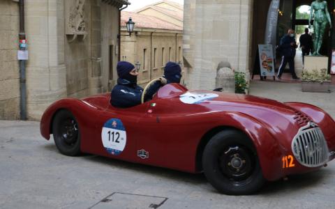 Auto d'epoca rossa con due persone mascherate in un contesto urbano.